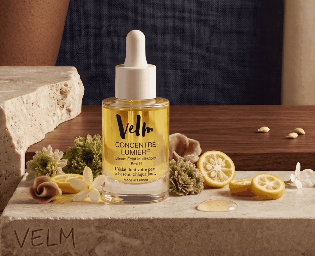 Un flacon compte-gouttes transparent contenant un sérum jaune doré, nommé « CONCENTRÉ LUMIÈRE » par Velm, est disposé au centre d'un décor graphique mêlant blocs de pierre et surfaces en bois. Il est entouré de tranches de petits agrumes jaunes, de fleurs miniatures et de boutons floraux séchés.