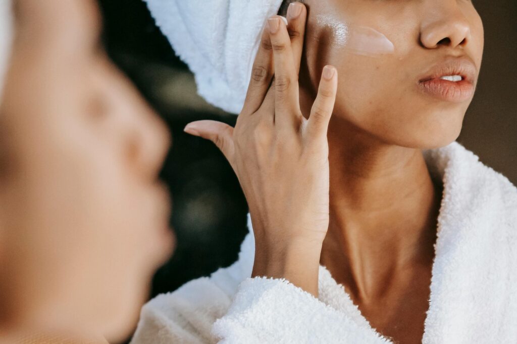 Une personne à la peau noire, vêtue d'un peignoir blanc et les cheveux enveloppés dans une serviette assortie, applique délicatement une crème de soin sur sa joue gauche. Son visage, de profil, montre une peau lumineuse et hydratée. On distingue également son reflet flou dans un miroir situé au premier plan à gauche.