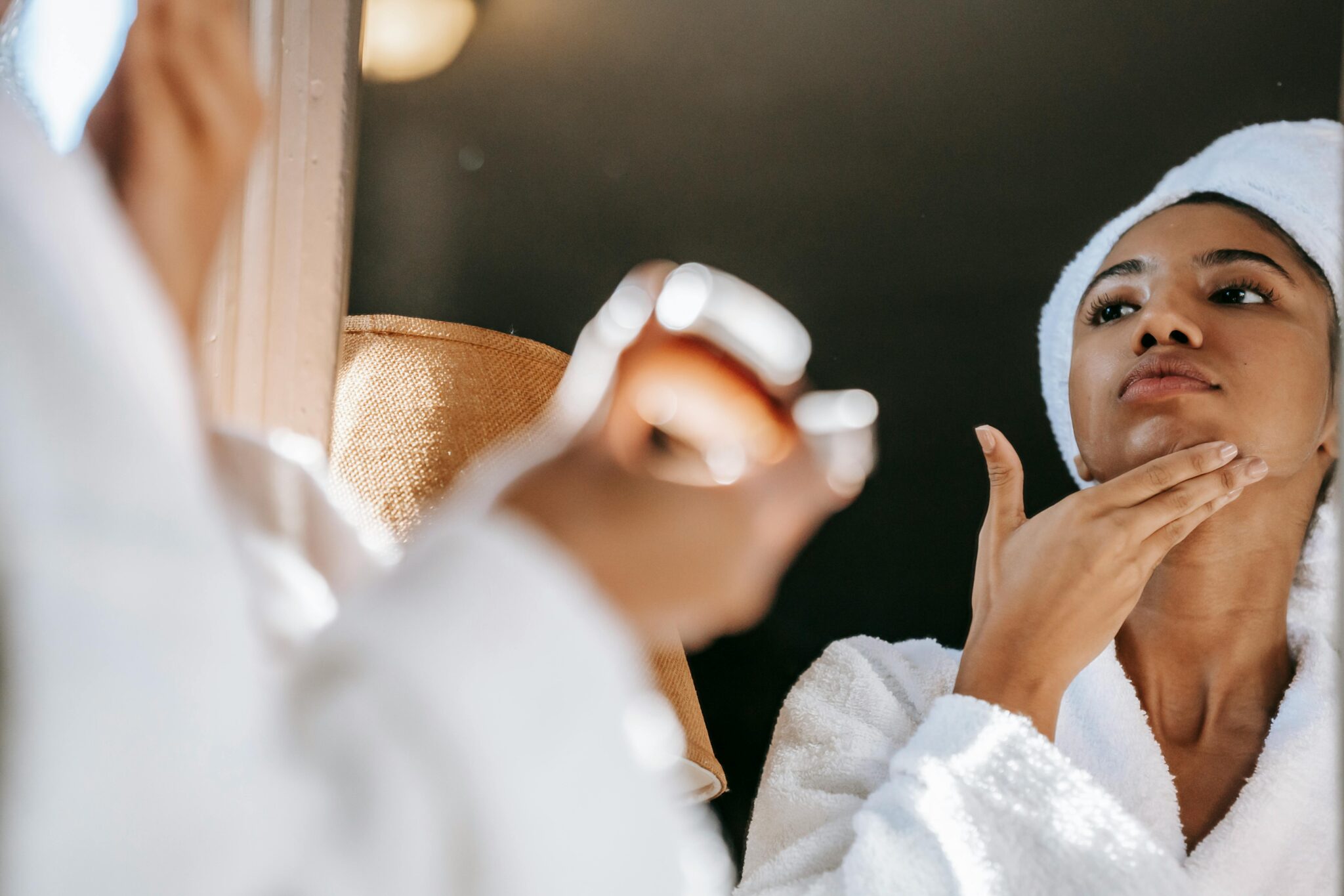 Femme aux cheveux attachés, en peignoir blanc, s'appliquant de la crème sur le cou devant le miroir d'une salle de bain lumineuse