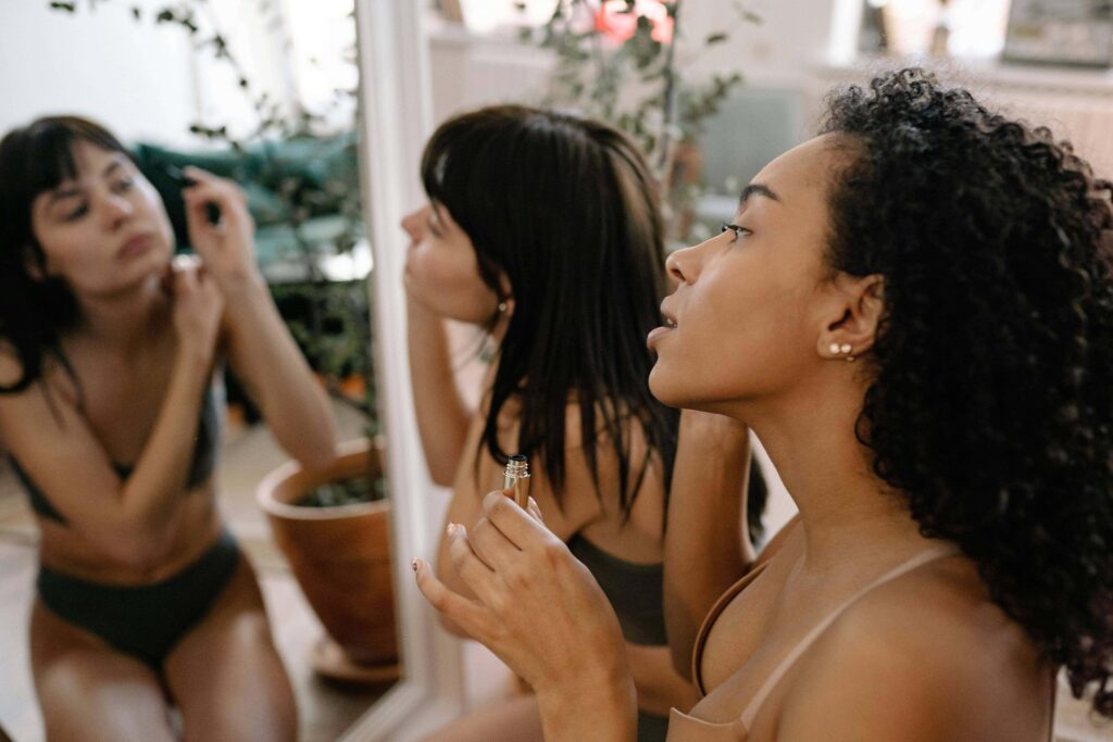 Deux jeunes femmes se préparent ensemble devant un grand miroir. Au premier plan, une femme noire aux cheveux bouclés, vue de profil en sous-vêtements beiges, tient un applicateur de maquillage. Derrière elle, une femme aux cheveux noirs et raides se maquille également, son reflet ainsi que celui d'une troisième personne étant visibles dans le miroir.