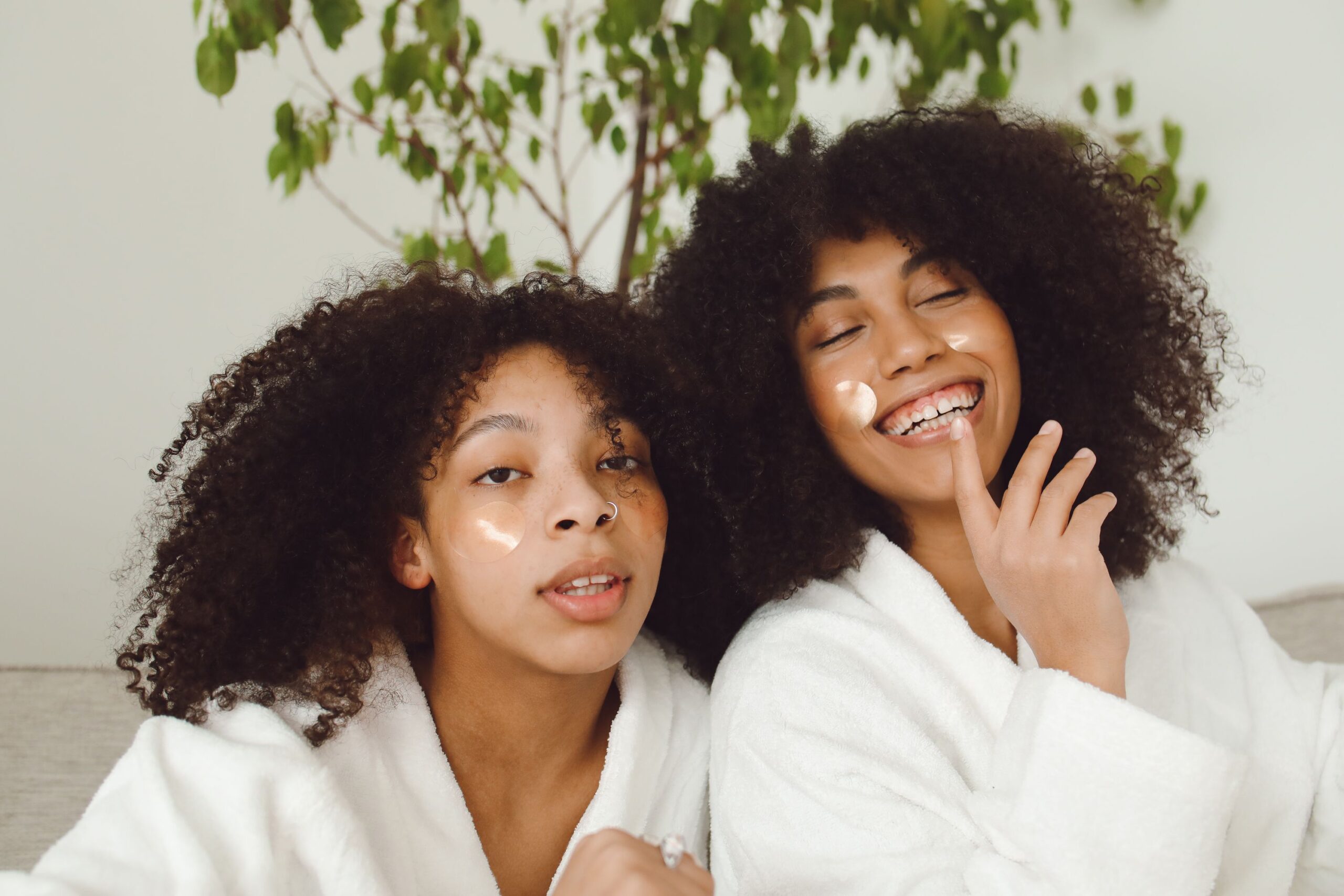 Deux amies à la peau métisse et cheveux crépus, en peignoir blanc, portant des patchs pour le contour des yeux et souriant.