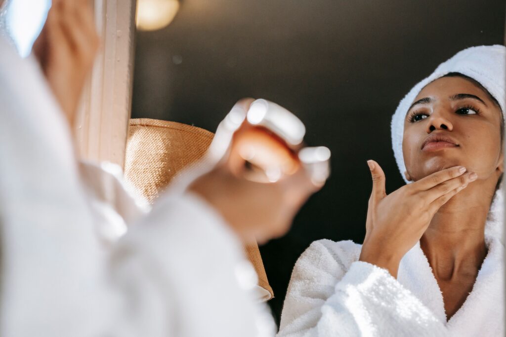Femme aux cheveux attachés, en peignoir blanc, s'appliquant de la crème sur le cou devant le miroir d'une salle de bain lumineuse